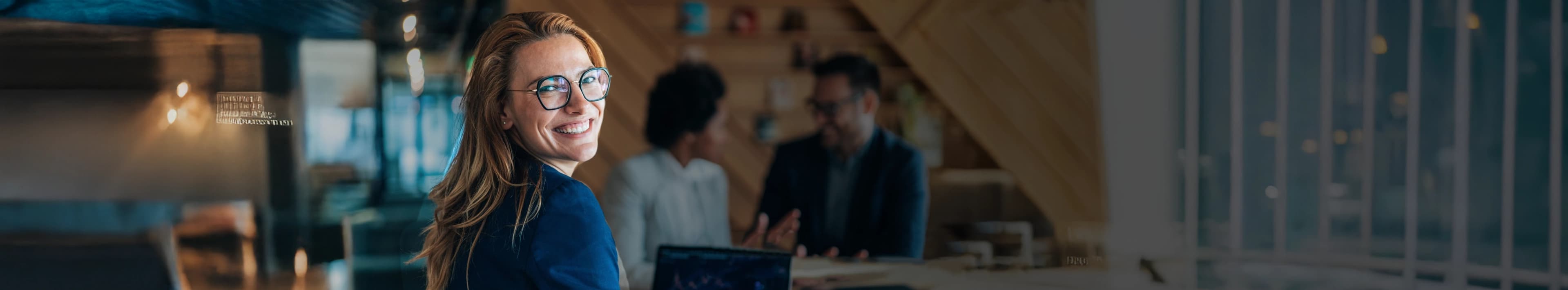 Eine lächelnde Geschäftsfrau blickt in die Kamera, während ihr Team im Hintergrund zusammenarbeitet.