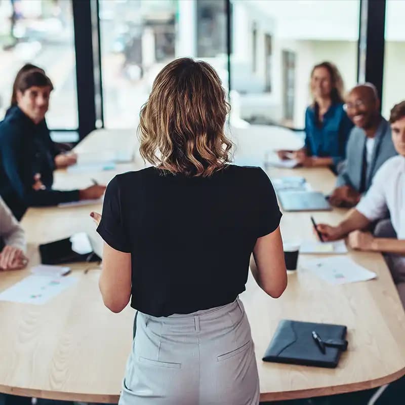 Eine Frau steht bei einem Meeting und präsentiert dem aufmerksamen Team vor ihr Ideen.